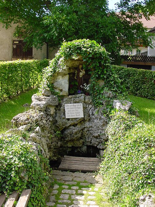Im Mittelpunkt der Antoniusbrunnen  umgeben von grünem Bewuchs im Hintergrund sind Häuser zu erkennnen.