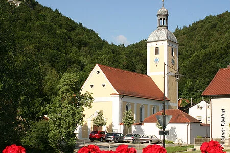 Die Kirche von Mühlbach von der Brücke aus, vorne  Geranien, im Hintergrund der bewaldete Hang.