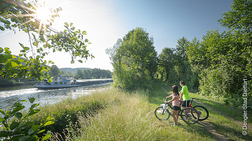 Zwei Radfahrer am Ufer des Rhein-Main-Donau-Kanal im Hintergrund ein Güterschiff auf dem Kanal