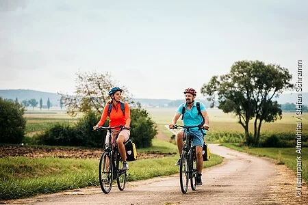 Eine Frau und ein Mann radeln auf dem Altmühltal-Radweg bei Dittenheim. Beide tragen einen Fahrradhelm und einen Rucksack. Der Radweg wird von Gras und grünen Bäumen gesäumt.