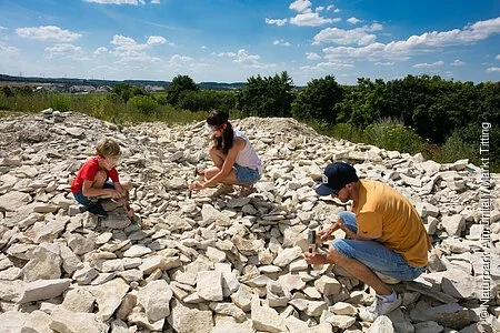 Eine Familie sucht an der Fossiliensammelstelle nach Versteinerungen. Alle drei knien mit Hammer und Meißel und versuchen die Platten zu teilen.