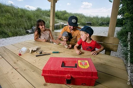 Eine Familie sitzt an einem Tisch an der Fossiliensammelstelle. Der Werkzeugkoffer mit Hammer und Meißel liegt vor Ihnen auf dem Tisch.