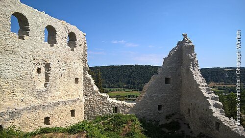 Links steht noch eine komplette Wand mit drei Rundbögenfenstern. Rechts sind nur noch die Teile zweier Mauern an der Ecke zu sehen. Im Hintergrund hat man einen schönen Blick ins Tal. Der Himmel ist wolkenlos.