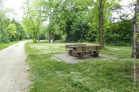 Unter dem Bäumen steht eine gemütliche Sitzgelegenheit aus Holz. Links führt ein geschotterter Weg entlang.