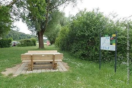Die Sitzgelegenheit aus Holz auf gepflastertem Grund ist inmitten einer grünen Wiese am Uferrand mit Büschen und Bäumen. Daneben steht eine Infotafel zum Thema Bootwandern.