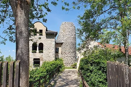 Der Eingang zur Burg Altmannstein ist mit einem Holzgeländer eingesäumt. Der weitere Weg ist mit Bäumen und Sträuchern auf beiden Seiten bepflanzt. Es ist der Turm und rechts ein Teil des Wohngebäudes zu erkennen.