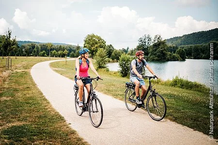 Eine Frau mit roten Shirt und blauem Fahrradhelm radelt neben einem Mann mit grauem Shirt und roten Helm. Rechts von ihnne liegt der Kratzmühlsee. Der Radweg ist von grünen Wiesen gesäumt. Der Himmel ist an diesem Tag etwas bewölkt.