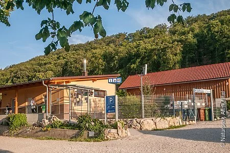 Das Gebäude aus Holz ist l-förmig gebaut und schön mit Pflanzen und Steinen angepflanzt. Hinter dem Campingplatz sieht man den Wald. Der Himmel strahlt himmelblau.