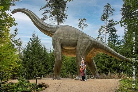 Eine vierköpfige, junge Familie bestaunt einen Dinosaurier aus Kunststoff, in voller Lebensgröße, auf einem Rundgang auf dem Besucherweg im Wald des Dinosauriermuseum Denkendorf.