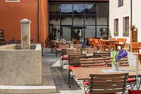 Der Biergarten der Gastwirtschaft Schattenhofer  ist umgeben von den Wänden und dem Neubau im Hintergrund aus Glas. Links ist ein Steinbrunnen zu sehen.