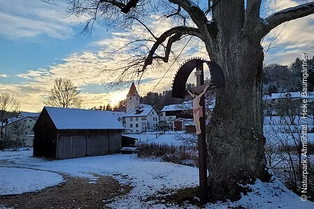 Winterweg mit Baum und einem Kreuz. Im Hintergrund Häuser und eine Kirche mit Schnee bedeckt.