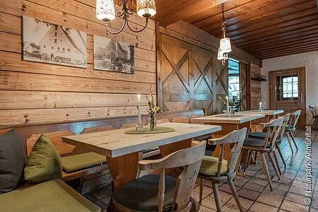 Modern gestalteter Teil der Gaststube in der Krone in Kinding. Verwendung von Holz vereint Tradition und Moderne zum Beispiel durch helle, naturbelassene Holzwände, einer Holzdecke, eine Schiebetüre im Scheunenstil sowie helle Sitzmöglichkeiten mit Sitzkissen in gedeckten Farben.