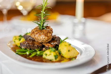 Das Bild zeigt einen Braten, welcher mit viel Hingebung angerichtet wurde. Drappiert ist der Braten auf einem Bett aus grünen Bohnen mit frischen Petersilien-Kartoffeln. Oben auf ein Semmelknödel garniert mit einem Rosmarin-Zweig und schöner Bratensoße