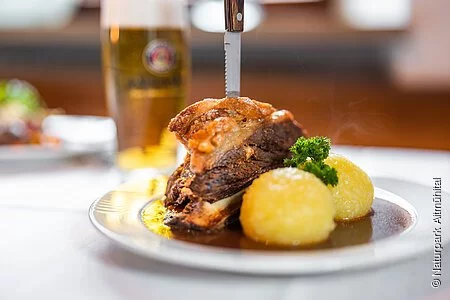 Traditionelles Schweineschäuferl, angerichtet auf einem weißen Teller mit viel Soße, zwei Kartoffelknödel und einem scharfen Messer, welches traditionell senkrecht in das Schäuferl gesteckt wird