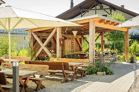 Ein liebevoll und grün gestalteter Biergarten mit Holz-Biertischen, großen weißen Sonnenschirmen sowie einem massiven Holzunterstand, der mit alten Küchenutensilien und Lichterketten dekoriert ist