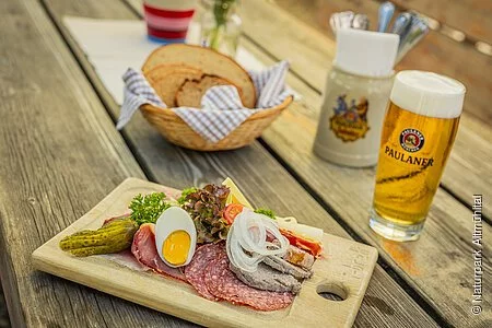 Auf einem Holz-Biertisch steht ein frisch gezapftes Glas helles Bier, ein Brotkorb mit bayerischer Serviette und frischem Brot drin sowie eine liebevoll und typisch bayerische Brotzeitplatte mit frischer Wurst, garniert mit Zwiebeln, Essiggurken und Ei