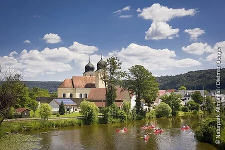 Auf der Altmühl paddeln fünf rote Kanus an Kottingwörth mit ihrer Kirche und deren zwei Kirchtürmen vorbei.