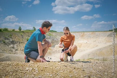 Ein Mann im blauen Shirt kniet neben einer Frau, die mit Hammer und Meißel vorsichtig eine Steinplatte im Fossiliensteinbruch Blumenberg teilt.