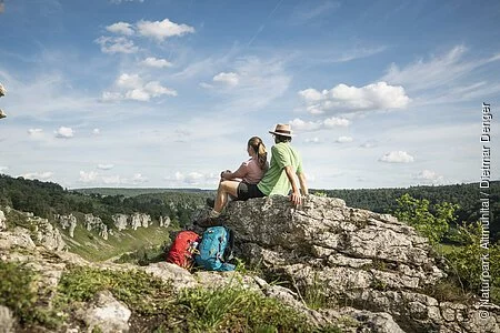 Ein Pärchen sitzt auf einem Felsen. Ihre Wanderrucksäcke lehnen daran. Sie lassen ihren Blick ins Tal schweifen.