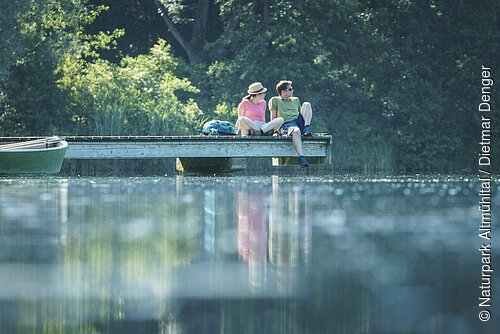 Ein Wanderpärchen sitzt auf dem Steg am Kratzmühlsee und lässt den Blick über den See schweifen. Hinter ihnen die Bäume und links am Steg ist ein Ruderboot befestigt.