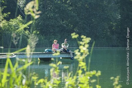 Ein Wanderpärchen sitzt auf dem Steg am Kratzmühlsee und lässt den Blick über den See schweifen. Hinter ihnen ragen die Bäume mit ihrem grünen Blätterkleid auf und links am Steg ist ein Ruderboot befestigt.