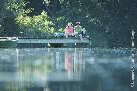 Ein Wanderpärchen sitzt auf dem Steg am Kratzmühlsee und lässt den Blick über den See schweifen. Hinter ihnen die Bäume und links am Steg ist ein Ruderboot befestigt.