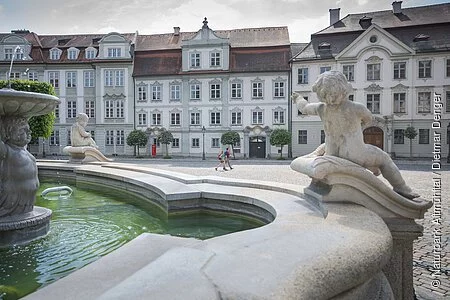 Ein Wanderpärchen ist auf dem Residenzplatz Richtung Brunnen unterwegs, dem man in Teilen sieht. Hinter ihnen reihen sich die historischen Bauten rund um den Platz auf.