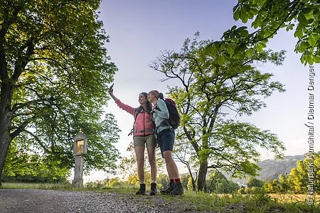 Zwei Frauen in rosafarbenen und hellblauem Shirt, sowie kurzen Hosen stehen eng beieinander am Frauenberg und die Frau in rosa, hält ihr Handy für ein gemeinsames Selfie in die Höhe. Ein großer Baum, sowie ein Marterl sind vor den Zweien zu erkennen. Im Hintergrund der zwei Frauen sind vereinzelt Bäume zu sehen , aber auch ein wolkenloser Himmel.