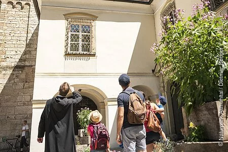 Eine Familie steht mit einem Mönch vor dem Eingang zum Kloster Plankstetten. Der Mönch erklärt der Familie etwas von der Geschichte des Gebäudes. Er hat seinen Arm gehoben und zeigt auf etwas.