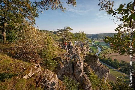 Ein Wanderpärchen, etwas entfernt, steht links auf einem Felsen an der Arnsberger Leite, die gut zu sehen ist. Dahinter erstreckt sich der Blick ins Tal. Der Himmel erstrahlt wolkenlos.