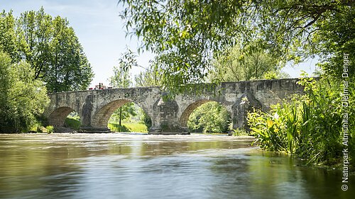 Auf der Römerbrücke sitzen zwei Wanderer Rücken an Rücken auf dem Steingeländer der Brücke. Eingerahmt von Bäumen und unter ihnen die Altmühl.
