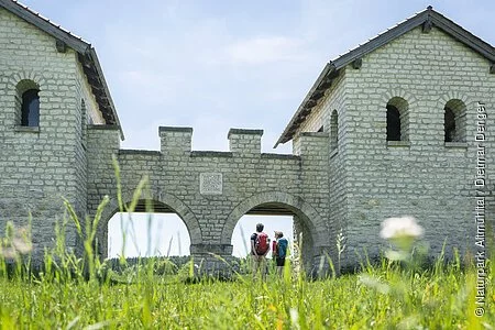 Zwei Wanderer stehen am Nordtor des Römerkastells. Das Kastell wurde rekonstruiert und mit hellen Steinen gebaut. Rechts und links sind zwei Türme mit Dächern, die mit zwei Rundbögen miteinander verbunden sind.