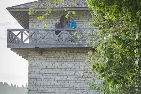 Eine Frau mit Strohhut und ein Mann mit blauer Jacke stehen an dem hölzernen Geländer oben auf dem Limesturm. Hinter ihnen ist eine hölzerne Tür mit der man ins innere des Turms gelangt. Der Turm hat weiter unten noch ein kleines Fenster und links sind einige Steinstufen. Rechts wird der Turm von grünen Blättern der Bäume eingerahmt.