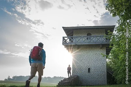 Ein Wanderer mit rotem Rucksack und blauer Jacke geht zum Limesturm. Auf den Treppen des Limesturm steht eine Frau und wartet. Der Limesturm ist komplett restauriert und ist aus weißem Stein gebaut. Oberhalb ist ein Geländer mit kleinem Rundgang aus Holz angebracht. Der Himmel ist bewölkt.