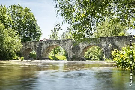 Auf der Römerbrücke sitzen zwei Wanderer Rücken an Rücken auf dem Steingeländer der Brücke. Eingerahmt von Bäumen und unter ihnen die Altmühl.
