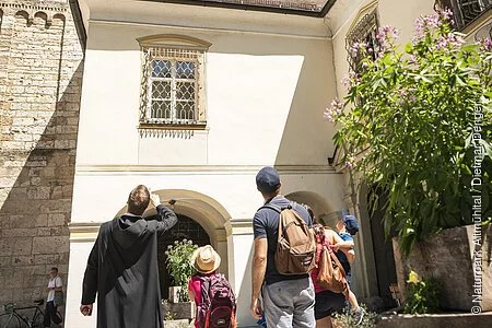 Eine Familie steht mit einem Mönch vor dem Eingang zum Kloster Plankstetten. Der Mönch erklärt der Familie etwas von der Geschichte des Gebäudes. Er hat seinen Arm gehoben und zeigt auf etwas.