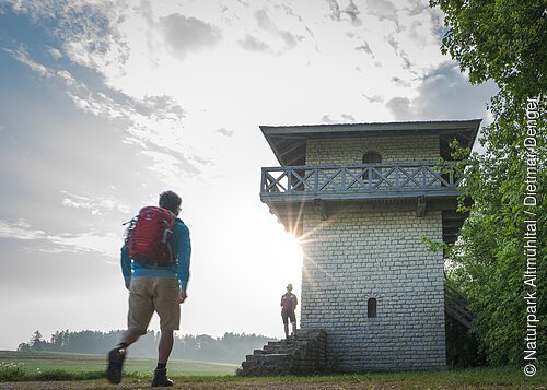 Ein Wanderer mit rotem Rucksack und blauer Jacke geht zum Limesturm. Auf den Treppen des Limesturm steht eine Frau und wartet. Der Limesturm ist komplett restauriert und ist aus weißem Stein gebaut. Oberhalb ist ein Geländer mit kleinem Rundgang aus Holz angebracht. Der Himmel ist bewölkt.