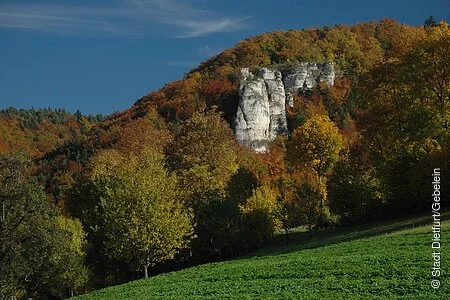 Der Kopffelsen in mitten von herbstlichen Laubbäumen, im Vordergrund ein Stück grüne Wiese und im Hintergrund blauer Himmel