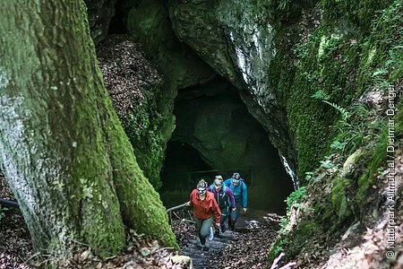 Aus dem Eingang der Höhle sind drei Wanderer nach oben unterwegs. Die Höhle liegt mitten im Wald und der felsige Eingang ist mit Moos übersät.