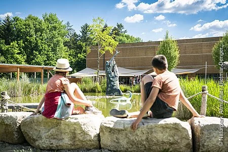 Zwei Kinder sitzen am Wasser im Außengelände des Dinosauriermuseum und beobachten die grüne Landschaft und das Wasser.