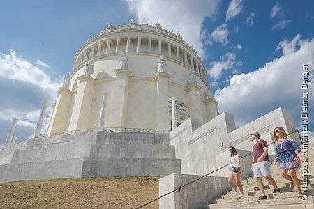 Zwei Frauen und ein Mann, in sommerlicher Kleidung, steigen die Treppen der Befreiungshalle hinunter und werfen einen letzten Blick zu ihr hinauf. Der Himmel strahlt blau und ein paar Wolken sind zu sehen.