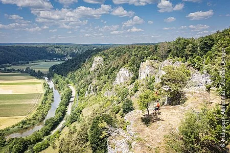 Sommerlicher Blick ins Altmühltal auf das Tal, auf den Fluss und auf die Felsenkette Arnsberger Leite, auf der ganz klein am Talrand eine Familile steht und ins Tal schaut.