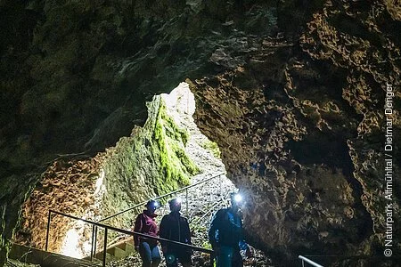 Drei Wanderer steigen die Treppenstufen zur Höhle hinab. Der Vorderste ist mit einer Stirnlampe ausgestattet und leuchtet ihnen den Weg aus. Licht kommt nur vom Eingang in die Höhle.