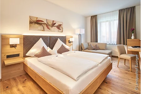 Ein großes Doppelbett steht in einem hellen Raum mit weißen Wänden und braunen Elementen. Über dem Bett hängt ein Bild.