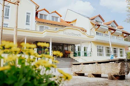 Der äußere Eingangsbereich des Hotels mit seinen Treppen und Säulen ist zu sehen. Im Vordergrund sind gelbfarbene Blumen und eine Brunnen zu sehen.