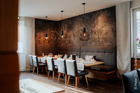 Es ist eine lange Tafel mit gepolsterten Stühlen und einer Eckbank aufgebaut. Im Hintergrund ist die Wand mit dunkelbraunen Ziertapeten festlich gestaltet.