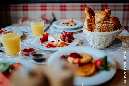 Der Frühstückstisch ist mit Orangensaft, Marmeladen, Pancakes und einem gut gefüllten Brötchenkorb gedeckt.