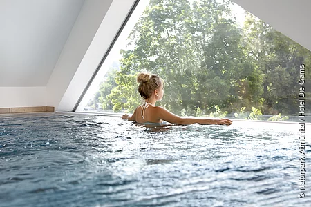 Ein Gast genießt den Ausblick von Schwimmbecken durch ein Fenster nach draußen in die Naturlandschaft.