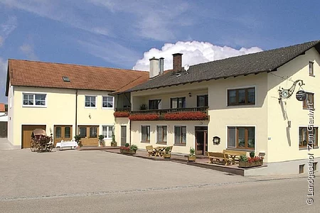 Der Landgasthof in L-Form ist hellgelb gestrichen. Der Balkon ist mit Geranien bestückt.