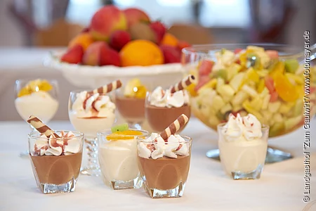 Lecker sechs Dessertgläser mit weißer und Schokoladenmousse stehen auf einem Tisch mit weißer Tischdecke. Im Hintergrund ist eine große Schale mit Obstsalat und eine Obstschale.